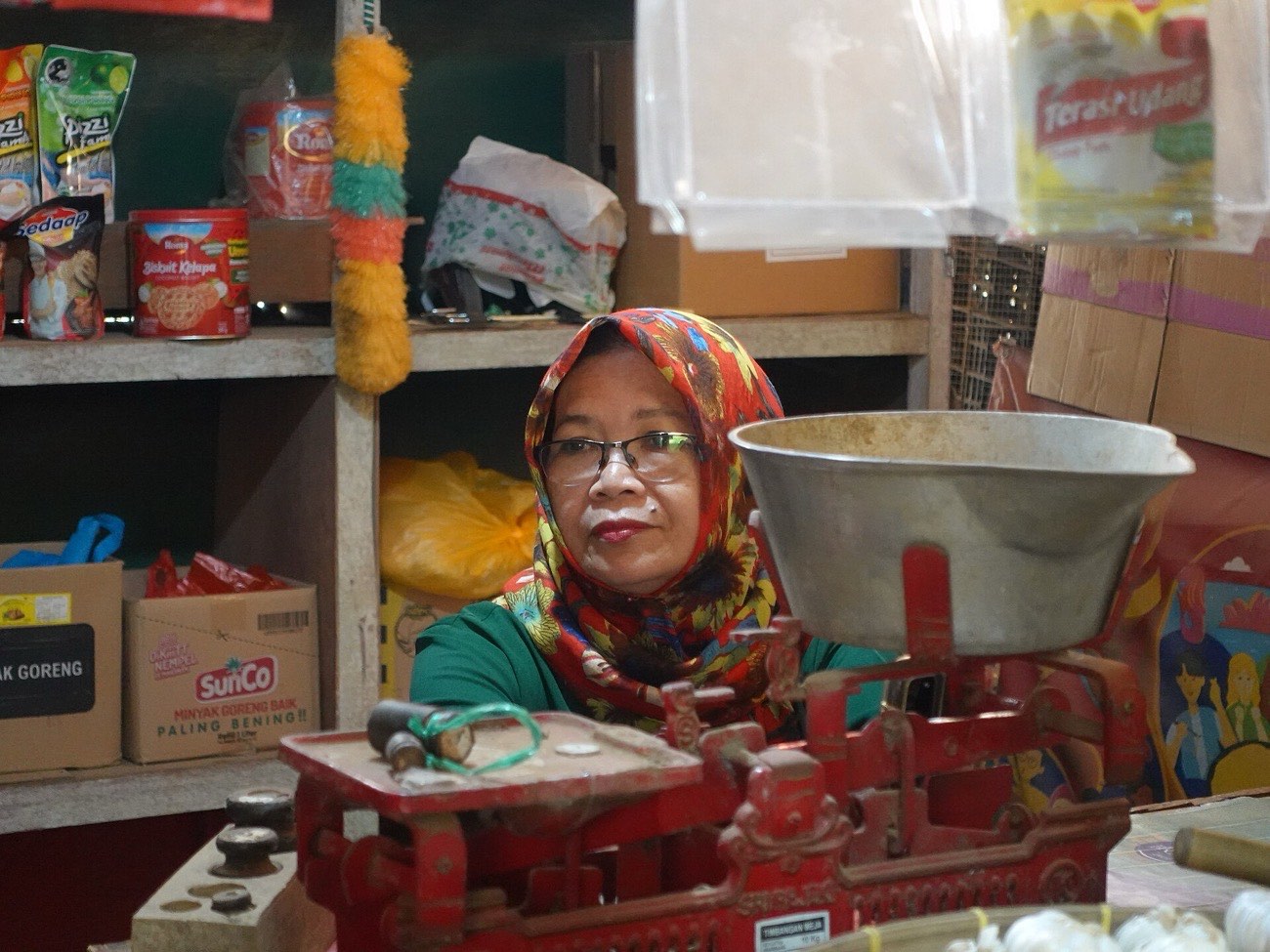 Lapak kebutuhan dapur dengan rak berisi bumbu, minyak, dan sembako, serta timbangan di depan